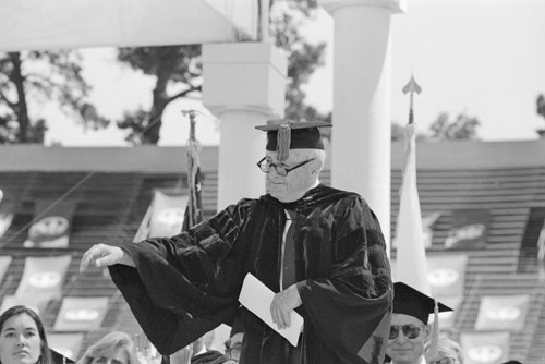 Photo of the late John W. Gardner in a cap and gown gesturing toward the crowd.