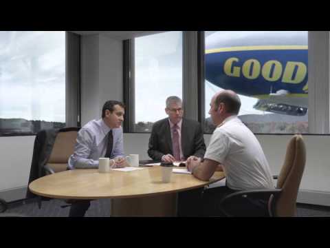 Three men sitting around a conference table. The Goodyear blimp is flying outside the window.