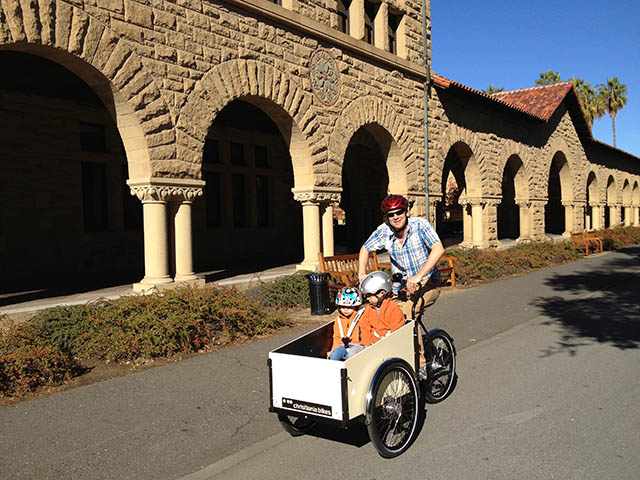 Photo of a dad biking with two small children in a box trailer.